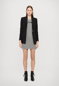 Blazer nero su un abito grigio a righe con il testo "Moschino Jeans". Modella indossa stivali neri alla caviglia. Sfondo semplice.