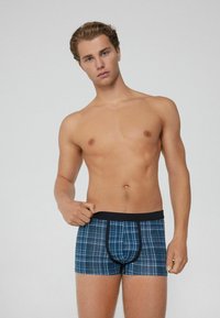 Muž bez trička v modrých kostkovaných trenýrkách, držící pásek jednou rukou proti jednoduchému světlému šedému pozadí.