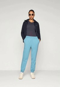 Šviesiai mėlynos sportinės kelnės su elastinga juosta, derinamos su tamsiai mėlyna zip-up striuke ir prigludusia juoda palaidine. Baltos sportinės bateliai ir akiniai nuo saulės.