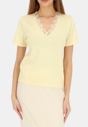 Kobieta ubrana w jasnożółtą bluzkę z krótkim rękawem i dekoltem w serek, ozdobioną koronką, w połączeniu z kremową spódnicą, ręce spoczywają luźno po bokach.