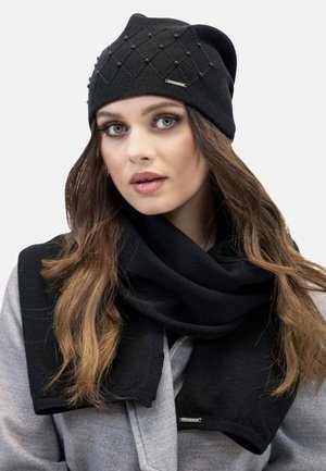 Zwarte gebreide beanie met een gestructureerd ruitpatroon en decoratieve kralen; gecombineerd met een zwarte sjaal. Beide items zijn gemaakt van zachte materialen.