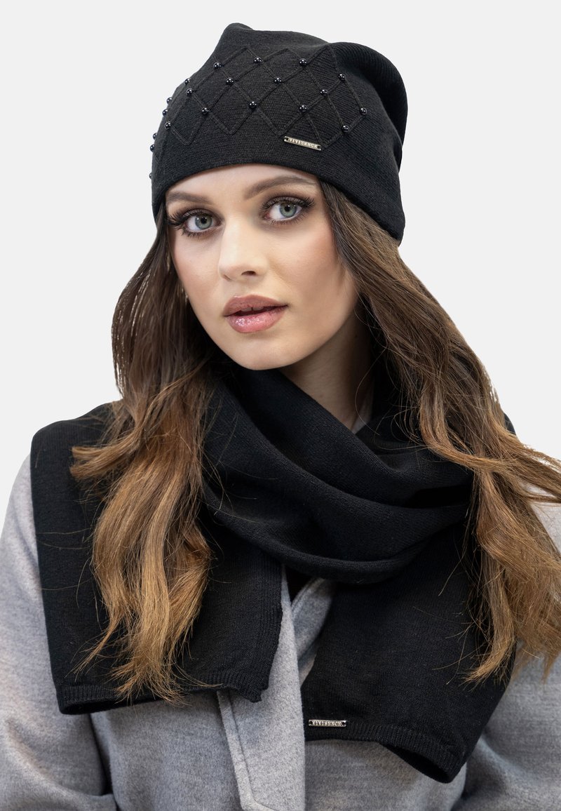Zwarte gebreide beanie met een gestructureerd ruitpatroon en decoratieve kralen; gecombineerd met een zwarte sjaal. Beide items zijn gemaakt van zachte materialen.
