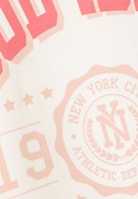 Camiseta de algodón blanca con gran impresión gráfica rosa que dice "NUEVA YORK" y un emblema circular, textura suave y estilo informal.