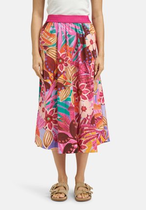 Woman wearing a colorful floral midi skirt with a pink waistband and beige buckle sandals, standing against a plain background.