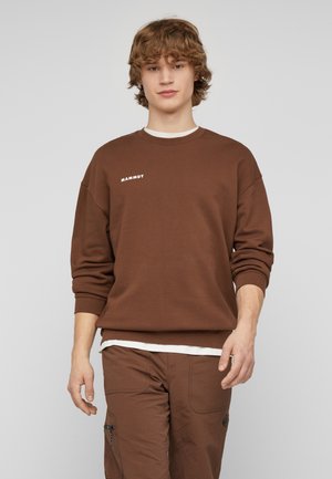 Giovane uomo con capelli ricci che indossa una felpa marrone Mammut e pantaloni abbinati su uno sfondo chiaro uniforme.
