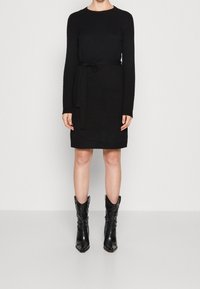 Abito in maglia nera con scollatura rotonda, maniche lunghe e cintura in tessuto. Indossato con stivali cowboy neri a punta, con una finitura testurizzata.