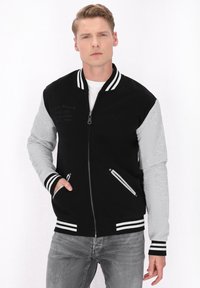 Jachetă varsity neagră și gri cu manșete și guler rayé, fermoar pe față și buzunare laterale. Prezintă un design texturat pe partea din față.