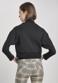 Fekete, rövidített pulóver lekerekített nyakkivágással és bordás mandzsettákkal, sárga akcentussal ellátott kockás nadrággal párosítva, amely hátul zsebekkel rendelkezik.