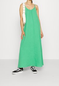 Zielona sukienka maxi z lekkiego materiału, z cienkimi ramiączkami i luźnym fasonem, zestawiona z czarnymi tenisówkami.