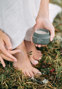Grön behållare märkt "Forest Barefoot Balm" håller en fot där en krämig balsam appliceras, placerad på mossa och tallnålar.