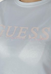 Lichtblauw shirt met korte mouwen van gladde stof, met grote, zachte roze letters van "GUESS Los Angeles CA" op de voorkant.