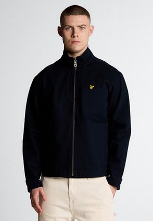 Giacca zip-up navy con colletto alto, tasche laterali e un piccolo logo giallo sul petto. Tessuto liscio con una vestibilità sartoriale.