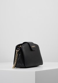 Borsa in pelle nera DKNY con chiusura a pattina e tracolla in catena dorata, posizionata su un piedistallo bianco contro uno sfondo grigio uniforme.