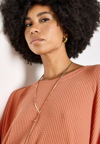 Top de punto texturizado color durazno con un escote ancho; lleva un collar largo y ondulado de oro y pequeños pendientes de aro dorados.