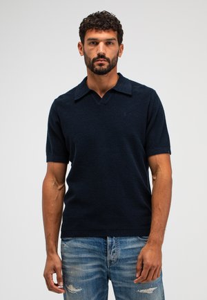 Man met kort krullend haar en baard, gekleed in een marineblauw gebreid shirt met korte mouwen en versleten blauwe spijkerbroek, staand tegen een effen achtergrond.