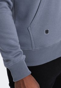 Sweatshirt gris avec des poignets et un ourlet côtelés, arborant une petite étiquette logo. Texture de tissu lisse, coupe décontractée et détail de poche latérale.
