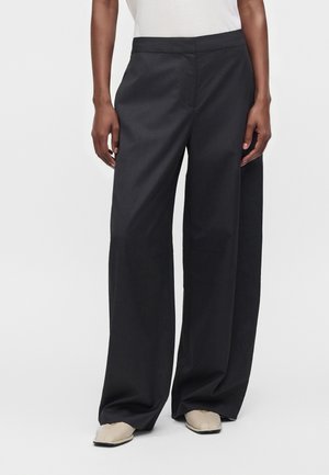Personne portant un pantalon noir taille haute à jambes larges et des chaussures pointues beige, debout devant un fond clair uni.