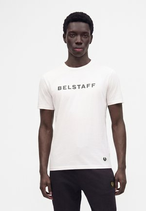 Mann mit kurzen lockigen Haaren, der ein weißes Belstaff-T-Shirt und schwarze Hose trägt, steht vor einem schlichten hellen Hintergrund.