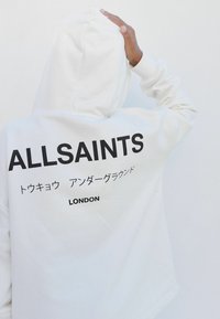 Bijela dukserica s kapuljačom na leđima s natpisom "ALLSAINTS" u podebljanom crnom tekstu, uz dodatni tekst na japanskom i "LONDON" ispod.