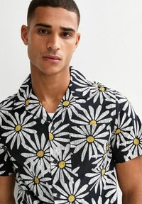 Chemise noire à manches courtes avec un motif floral de marguerites blanches et un centre jaune, dotée d'un col camp et d'une fermeture par bouton. Tissu texturé.