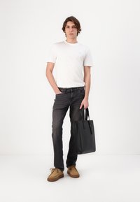Sac fourre-tout noir avec des bretelles verticales, porté par un mannequin portant un t-shirt blanc, un jean foncé et des chaussures beiges avec une semelle texturée.