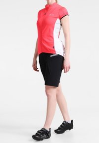 Różowa koszulka kolarska z krótkim rękawem i białymi bocznymi panelami, zestawiona z czarnymi szortami oraz czarnymi butami kolarskimi z regulowanymi paskami.