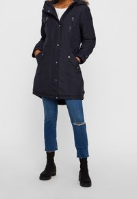 Person med knälång svart huva parka, vit skjorta, trasiga blå jeans och svarta ankelstövlar, stående med händerna i fickorna.