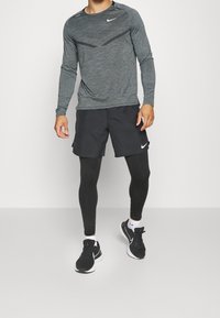Mann mit grauem langärmligen Sportshirt, schwarzen Shorts über schwarzen Leggings, weißen Socken und schwarzen Laufschuhen vor weißem Hintergrund.