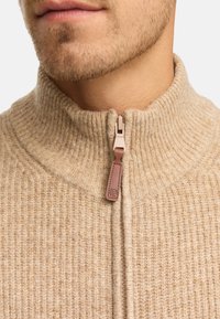 Beżowy sweter z ribbowanej wełny z wysokim kołnierzem i pół zamkiem. Zamek błyskawiczny ma brązowy skórzany uchwyt, co dodaje fakturalnego akcentu do projektu.