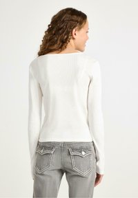 Langærmet hvid ribbet top med rundet halsudskæring, båret med lysegrå jeans med syninger og dekorative baglommer.