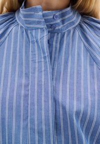 Ljusblå randig skjorta med hög krage, som har vertikala vita ränder och lila knappar, tillverkad av mjukt bomullstyg.