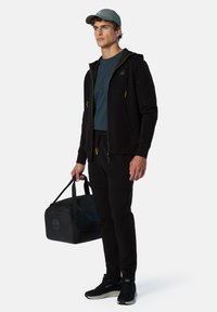 Felpa nera con zip e dettagli verdi, t-shirt scura, pantaloni tuta neri, sneakers neri e borsa a tracolla nera. Cappellino grigio indossato.