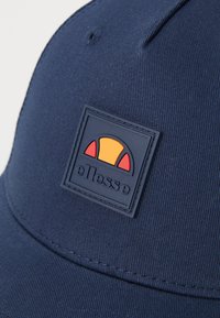 Sötétkék sapka, textúrált anyaggal, ívelt karimával és téglalap alakú logóval, narancssárga és piros részletekkel.