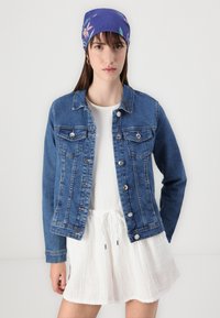 Jeans jakna u srednje plavoj boji s dugmadi i dva džepa na prsima. Nosena preko bijele haljine svezane konopcem. Ima ležeran, strukturiran dizajn.