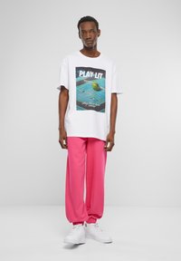 Hvit T-skjorte med en fargerik "PLAY LIT" grafikk av en tennisbane og en ball. Kombinert med løse rosa joggebukser og hvite joggesko.