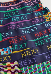 Grămadă de lenjerie colorată cu modele, cu talie elastică bleumarin pe care este inscriptionat cuvântul „NEXT” în diferite culori vii.