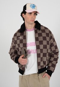 Homme portant une veste à carreaux beige et marron, chemise blanche avec texte rose, pantalon beige, casquette rose et beige, tenant des gants gris, regardant vers la droite.