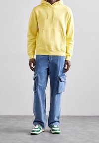 Felpa gialla con cappuccio e tasca frontale, abbinata a jeans cargo blu con due tasche laterali e sneakers con accenti verdi.