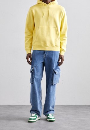 Sudadera amarilla con capucha y bolsillo frontal, combinada con jeans cargo azules que tienen dos bolsillos laterales y zapatillas con detalles en verde.