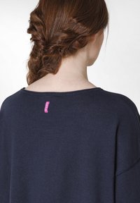 Marineblauer Pullover mit rundem Halsausschnitt und überschnittenen Schultern. Mit einem pinken Etikett am Nacken. Weiche Textur und bequeme Passform.