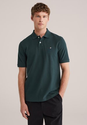 Jeune homme aux cheveux châtain clair portant un polo à manches courtes vert foncé et un pantalon noir, debout avec une main dans la poche.