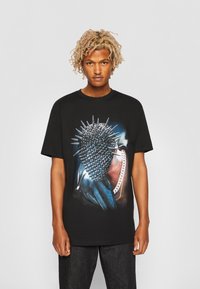Black cotton t-shirt featuring a graphic design of a spiked mask. The design incorporates metallic accents and a large chain detail.