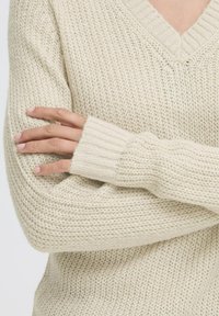 Beige gebreide trui met een V-hals, geribde mouwen en een losse pasvorm. De textuur is luchtig met een gematigd losse steekpatroon.