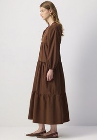 Bruine, lange mouw maxi-jurk met een gelaagd ontwerp en een gerimpelde zoom. Gemaakt van een soepele stof, gecombineerd met platte, bruine schoenen.