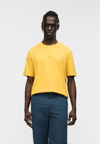 Κίτρινη βαμβακερή μπλούζα με κοντά μανίκια και στρογγυλό λαιμό, με διακριτικό λογότυπο στο στήθος. Συνδυάζεται με μπλε παντελόνια.