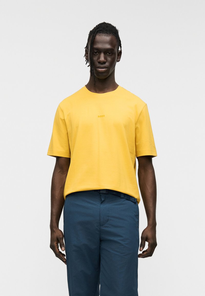 Κίτρινη βαμβακερή μπλούζα με κοντά μανίκια και στρογγυλό λαιμό, με διακριτικό λογότυπο στο στήθος. Συνδυάζεται με μπλε παντελόνια.