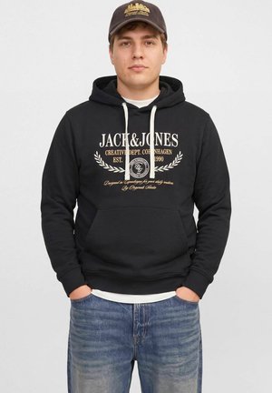 Un homme porte un sweat à capuche noir Jack & Jones avec des cordons blancs et une casquette marron. Il se tient debout, les mains dans les poches de son jean, sur fond blanc.