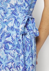 Gros plan d’un tissu à motif de feuilles tropicales bleu et blanc, noué en un nœud à la taille avec une partie d’un bras nu visible.