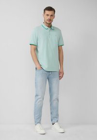 Polo shirt verde chiaro con colletto e polsini di un verde scuro in contrasto, indossato con jeans blu chiaro e sneakers bianche. Modello in posizione eretta.