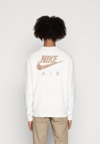 Vit sweatshirt med beige Nike Air-logotyp på ryggen. Har ribbade ärmslut och en avslappnad passform. Tillverkad av mjukt, strukturerat tyg.